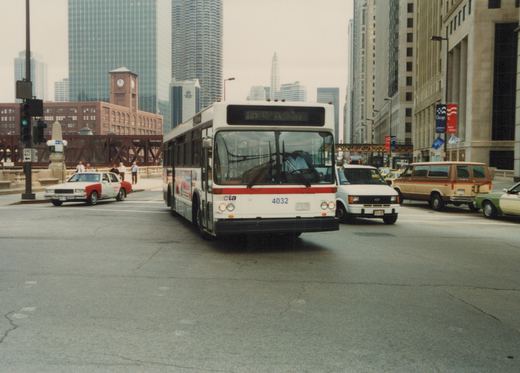 A white and red CTA bus with the number 4032 is stopped at a city intersection with tall buildings and other vehicles in the background.