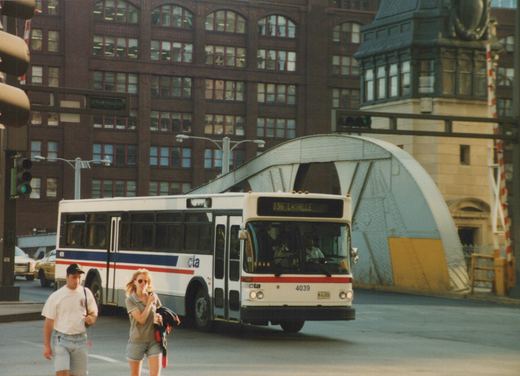 A white and blue Chicago Transit Authority bus, number 4039, is on a city street with a large bridge and pedestrians nearby.