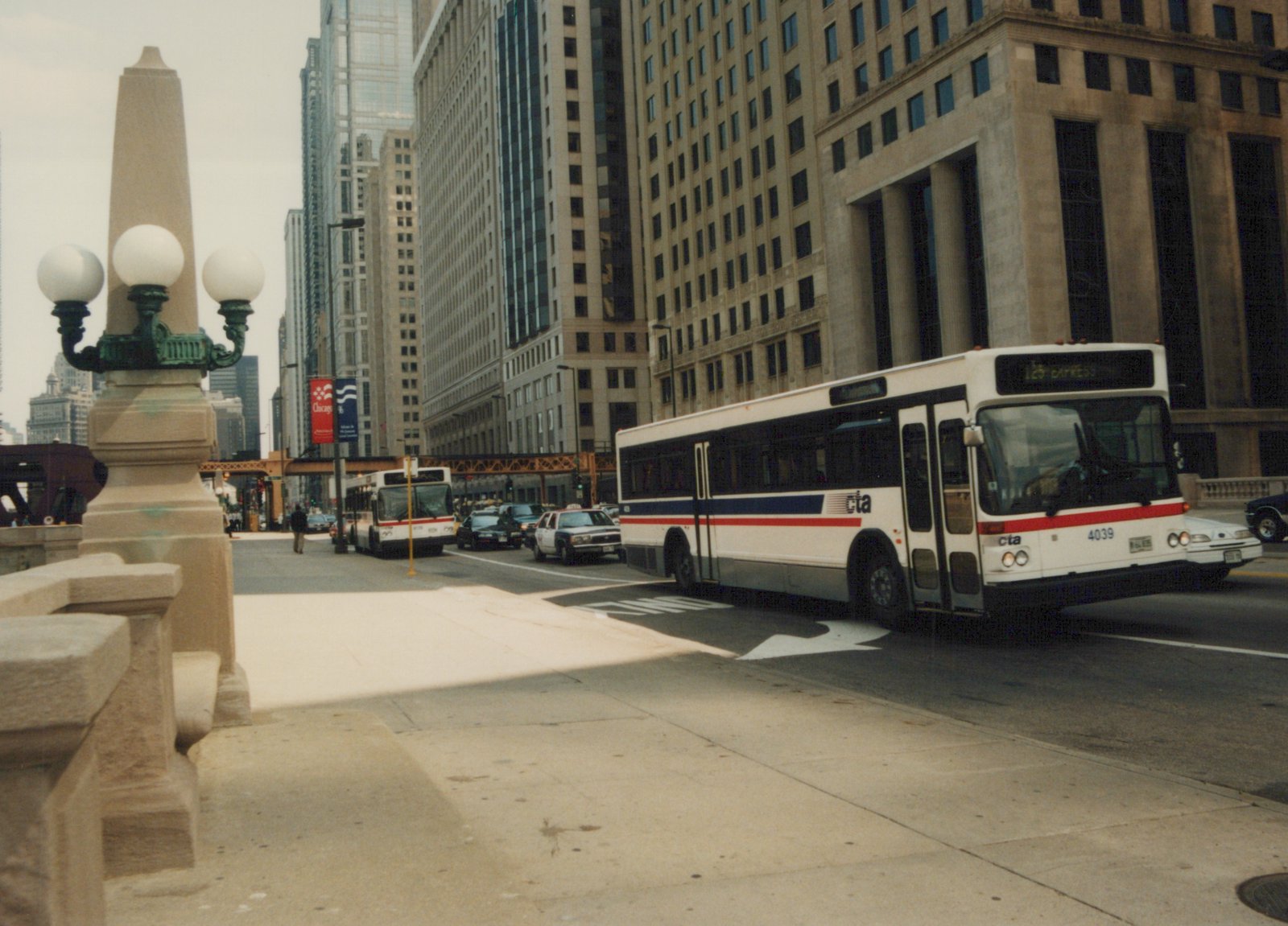 A white Chicago Transit Authority bus with a blue and red stripe, number 4039, is driving on a city street lined with tall buildings under an overcast sky.