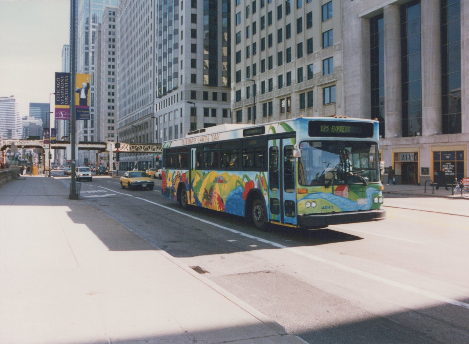 A colorful, Earth Day-themed Chicago Transit Authority bus, number 4041, is shown driving down a city street lined with tall buildings, with a few pedestrians and cars nearby.