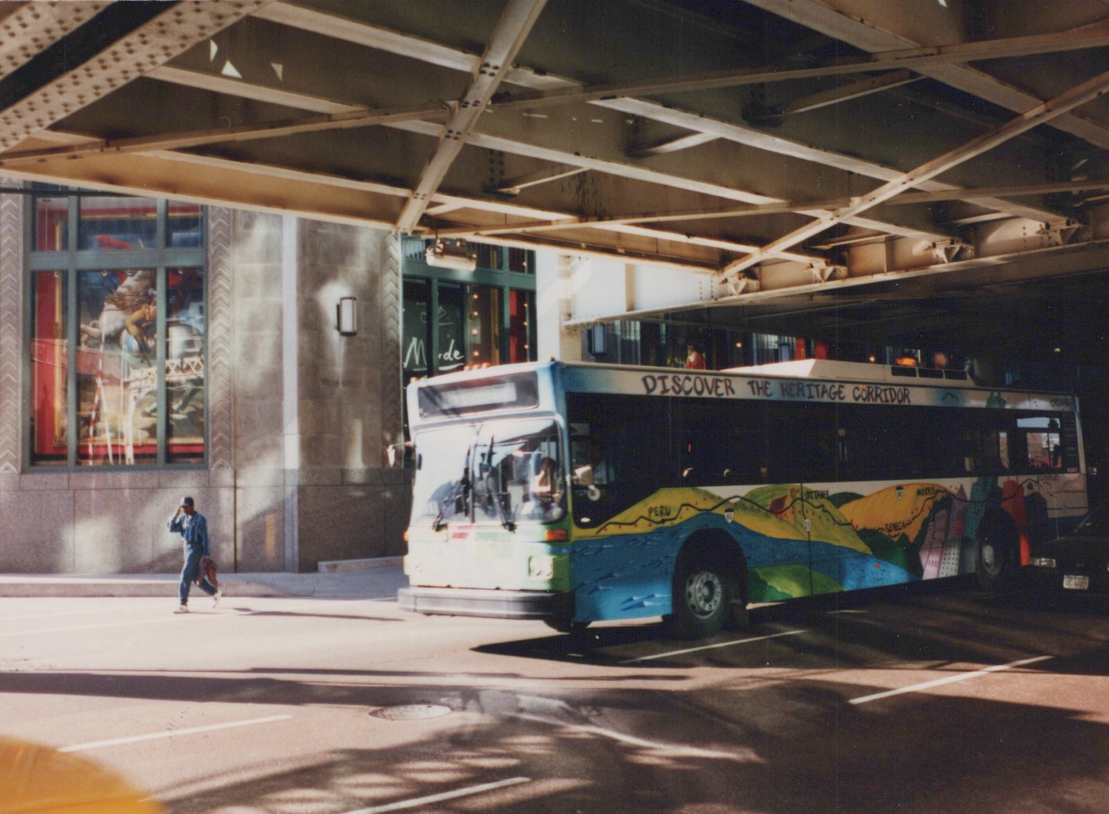 A white and blue motor bus with a colorful map painted on its side is stopped on a city street under a steel bridge on a sunny day, with a person walking on the sidewalk nearby.