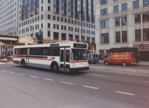 A white CTA bus with red and blue stripes, numbered 4044, drives down a city street with tall buildings and an elevated train track overhead. A red service van is parked on the side of the road.