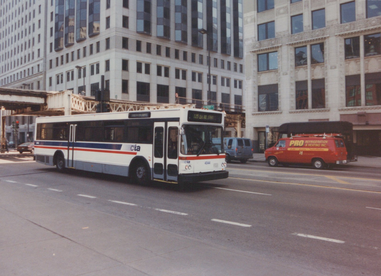 A white CTA bus with red and blue stripes, numbered 4044, drives down a city street with tall buildings and an elevated train track overhead. A red service van is parked on the side of the road.