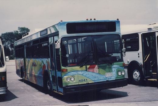 A Chicago Transit Authority bus with an Earth Day mural parked next to a white CTA bus, both on a paved lot on a sunny day.