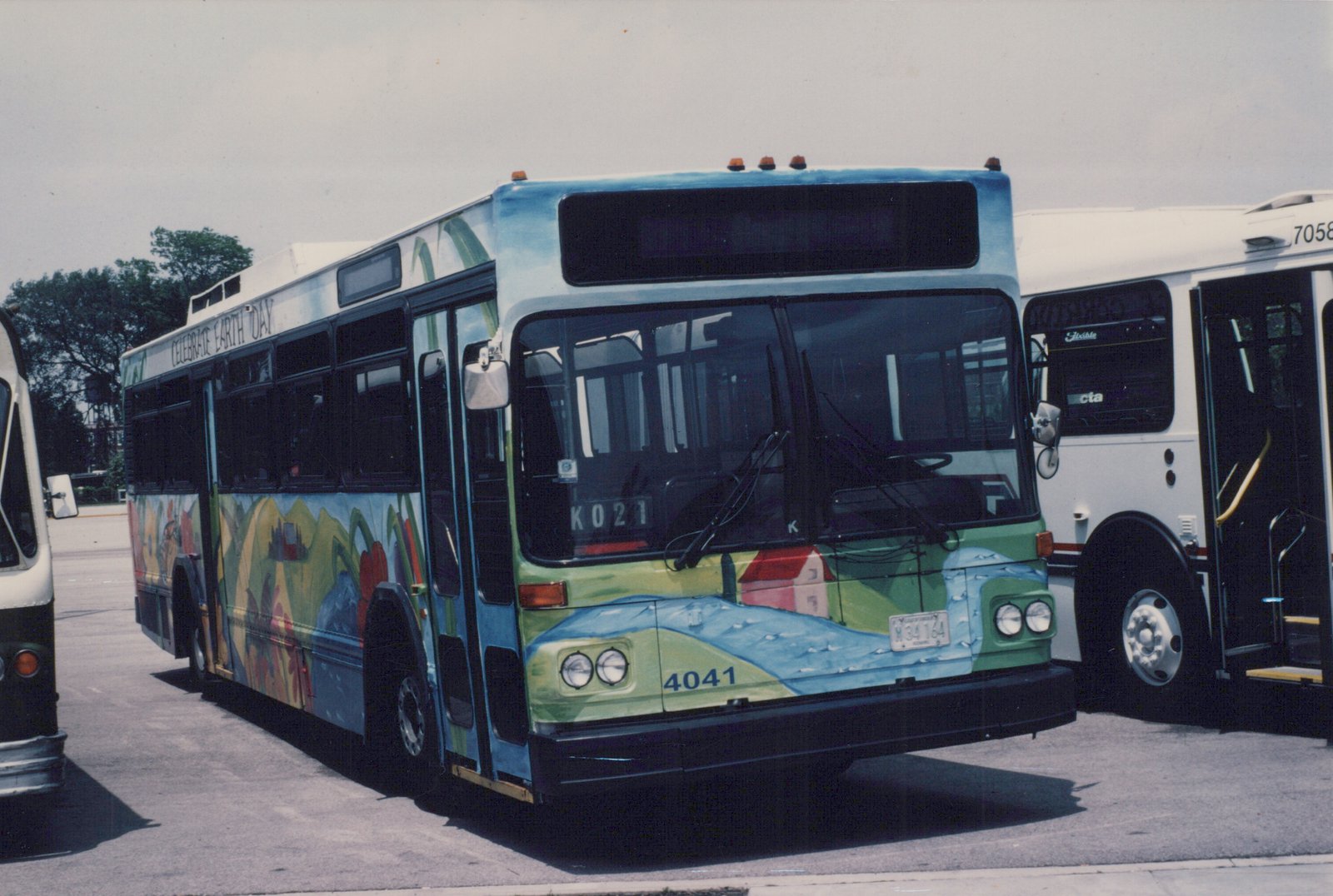 A Chicago Transit Authority bus with an Earth Day mural parked next to a white CTA bus, both on a paved lot on a sunny day.