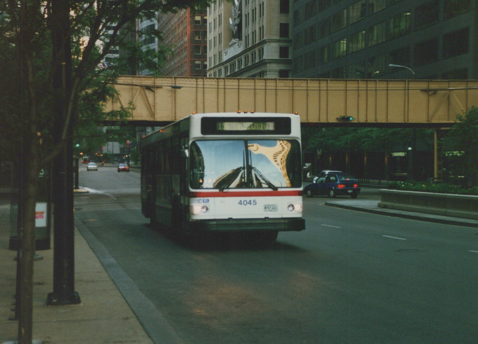 A white and red CTA bus with the number 4045 on the front, displaying route 156 to Belmont, travels down a city street with a yellow elevated railway structure overhead and tall buildings in the background.