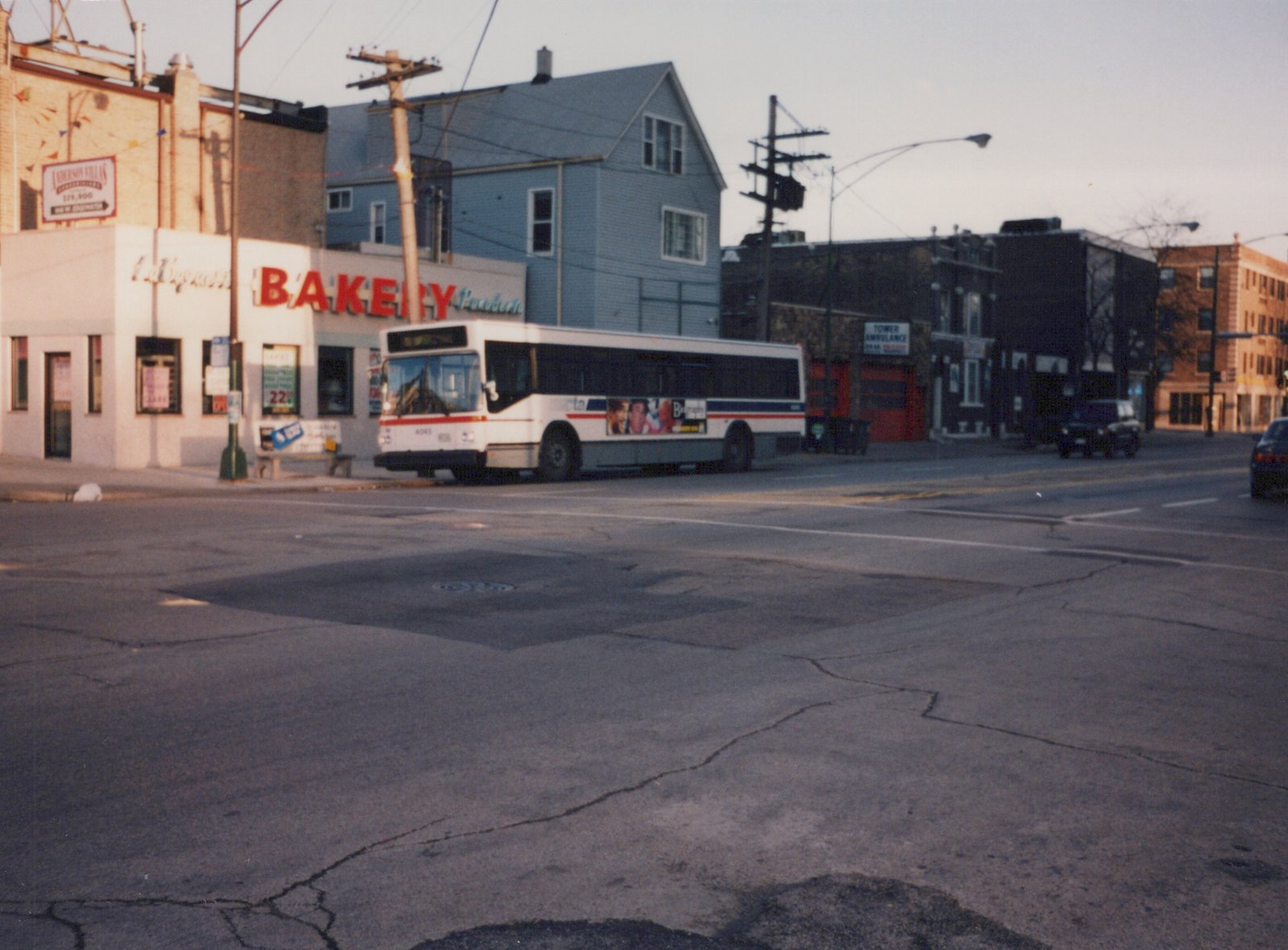 A white CTA bus with red and blue stripes, numbered 4045, is stopped on a city street at an intersection with a bakery and ambulance service visible in the background.