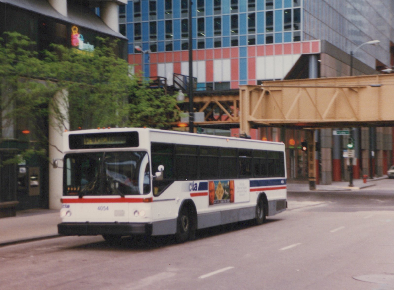 A white CTA bus with the number 4054 is driving on a city street, with a modern building and an elevated structure in the background.