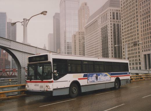 A white city bus with red and blue stripes is driving on a wet bridge in front of tall skyscrapers on a rainy day.