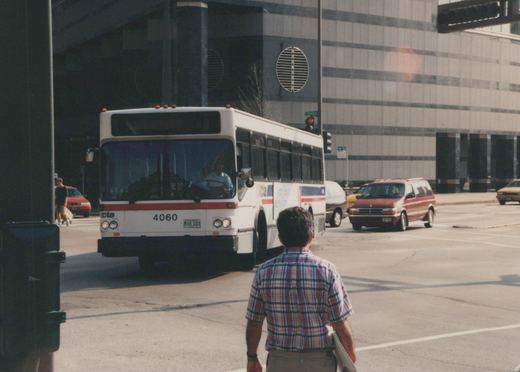A white and red Chicago Transit Authority bus, number 4060, is stopped at an urban intersection, with a man in a plaid shirt walking away from the camera in the foreground and other vehicles nearby.