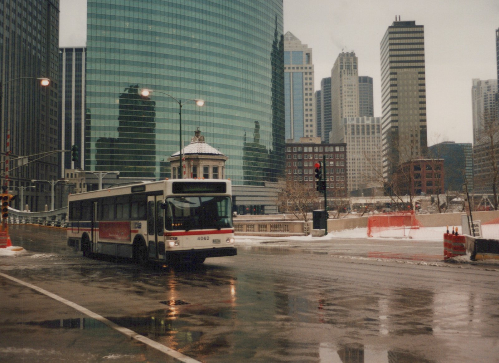 A white and red CTA bus drives on a wet, snow-dusted street in downtown Chicago, with glass skyscrapers and bare trees in the background.
