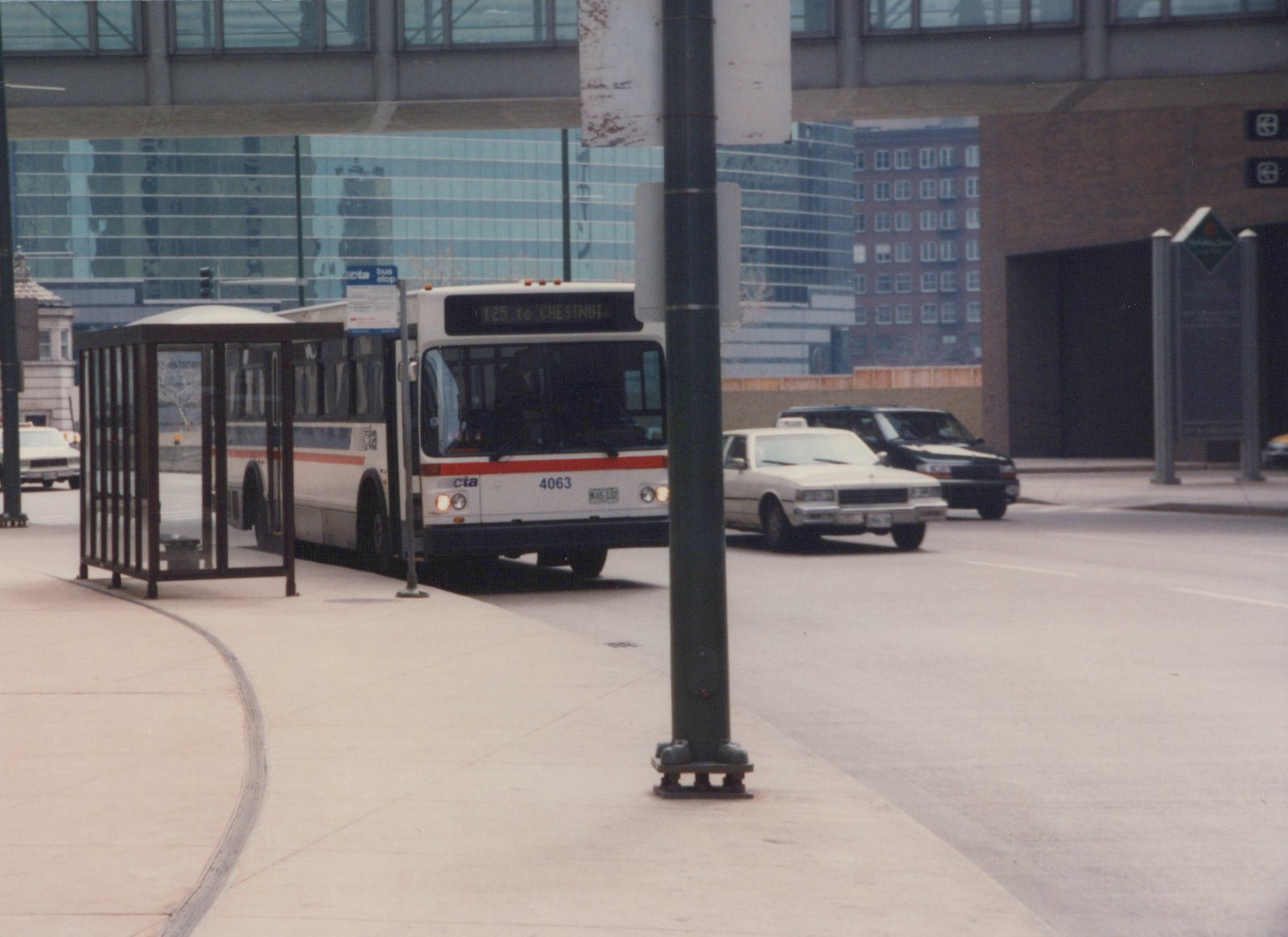 A white CTA bus with red and blue stripes, numbered 4063, is stopped at a bus shelter under an overpass in a city street; a taxi and another car are visible nearby.