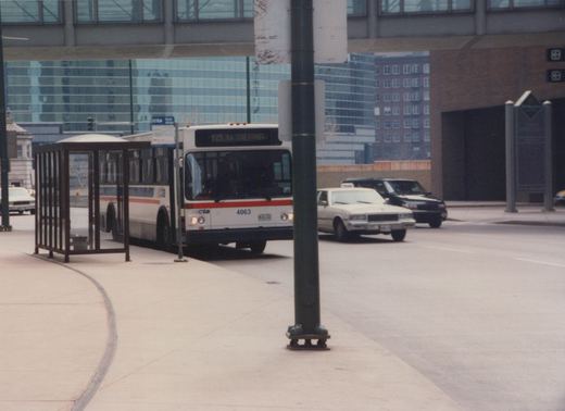 A white CTA bus with red and blue stripes, numbered 4063, is stopped at a bus shelter under an overpass in a city street; a taxi and another car are visible nearby.