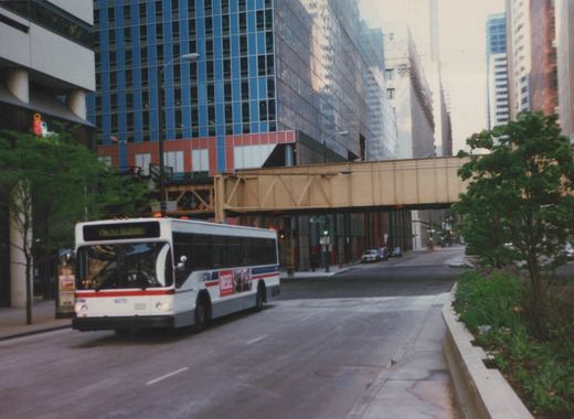 A white, red, and blue CTA bus with the number 4070 is driving on a city street under a brown steel elevated train structure, with tall modern buildings in the background.