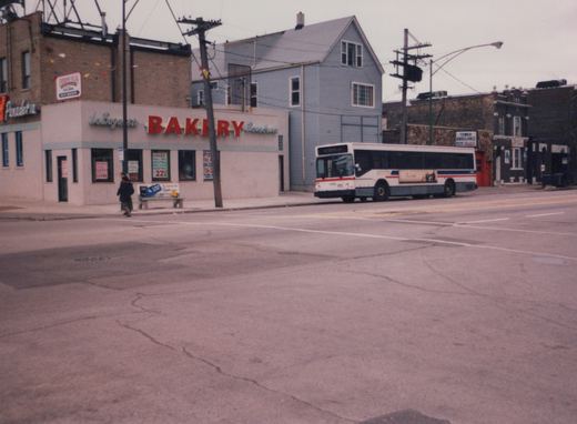 A white, red, and blue CTA bus numbered 4066 is parked on a street corner next to a bakery with a pedestrian crossing the road in the foreground.