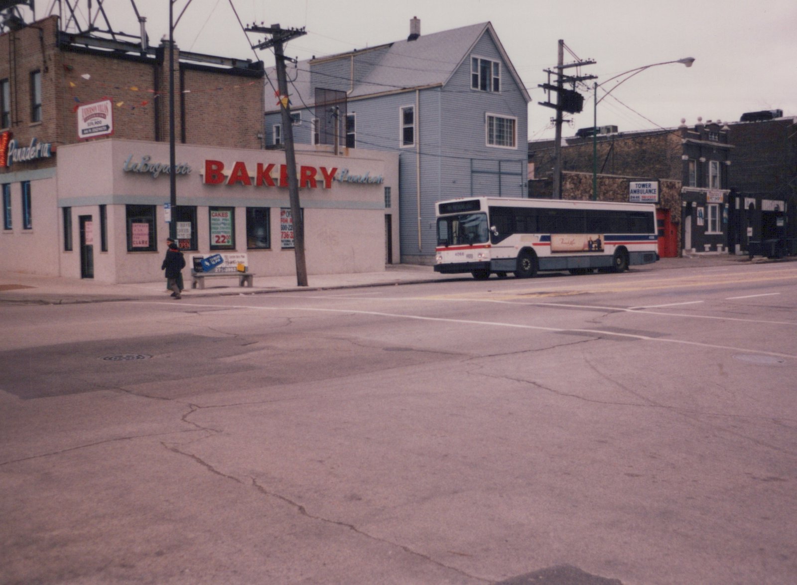 A white, red, and blue CTA bus numbered 4066 is parked on a street corner next to a bakery with a pedestrian crossing the road in the foreground.