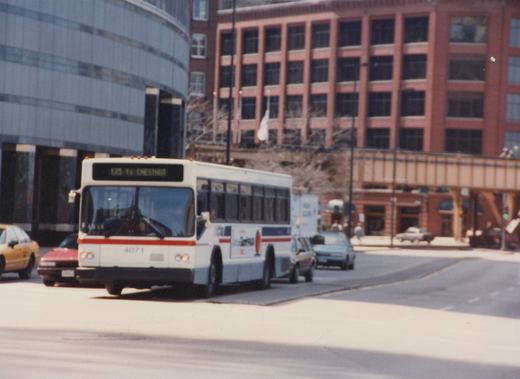 A white and red Chicago Transit Authority bus, number 4071, is stopped at an intersection with several cars and buildings in the background.