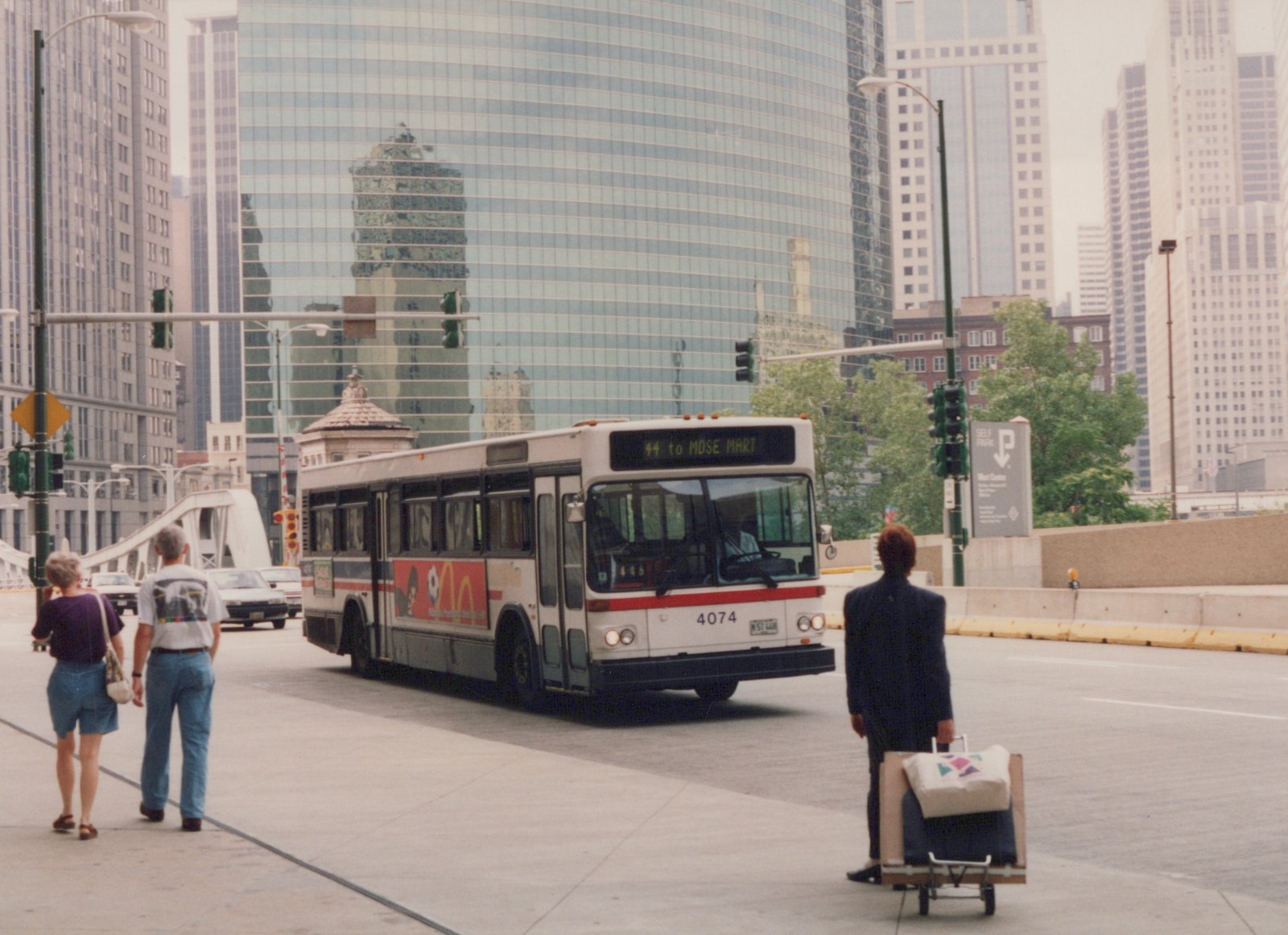 A white and red diesel bus with the number 4074 is driving on a city street with tall buildings in the background. Pedestrians and other cars are visible.