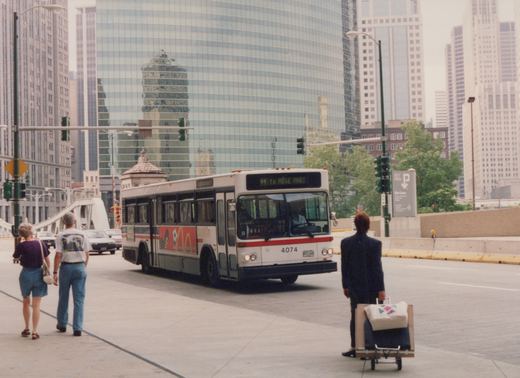 A white and red diesel bus with the number 4074 is driving on a city street with tall buildings in the background. Pedestrians and other cars are visible.