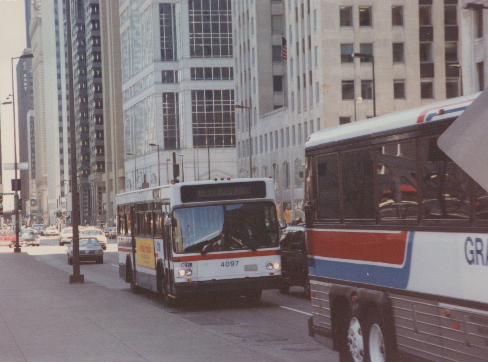 A white, red, and blue Chicago Transit Authority bus, number 4097, is on a city street with tall buildings in the background, and part of another large bus is visible to its right.