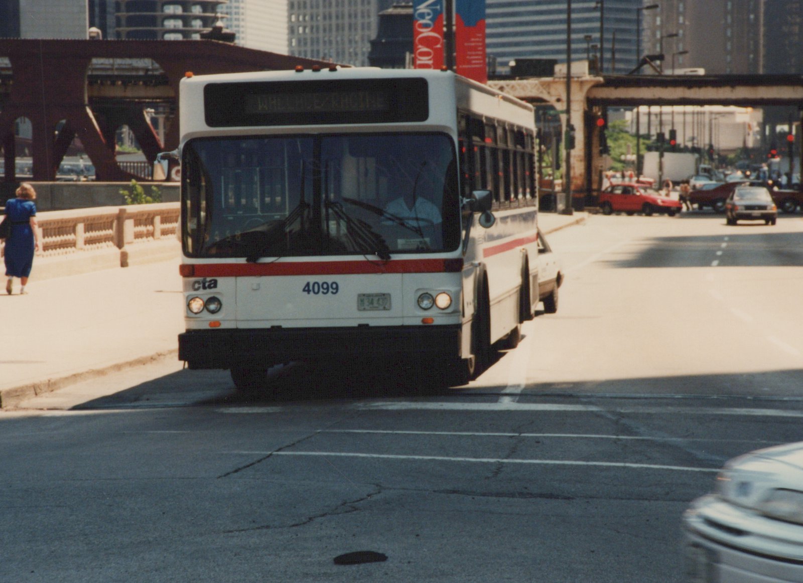 A white CTA bus with red stripe, number 4099, drives down a city street on a sunny day, with a bridge and some cars in the background.