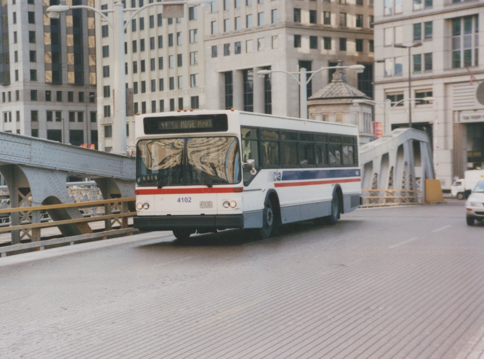 A white CTA bus with blue and red stripes, numbered 4102, displays its route as 44 to MUSE MART while crossing an urban bridge.