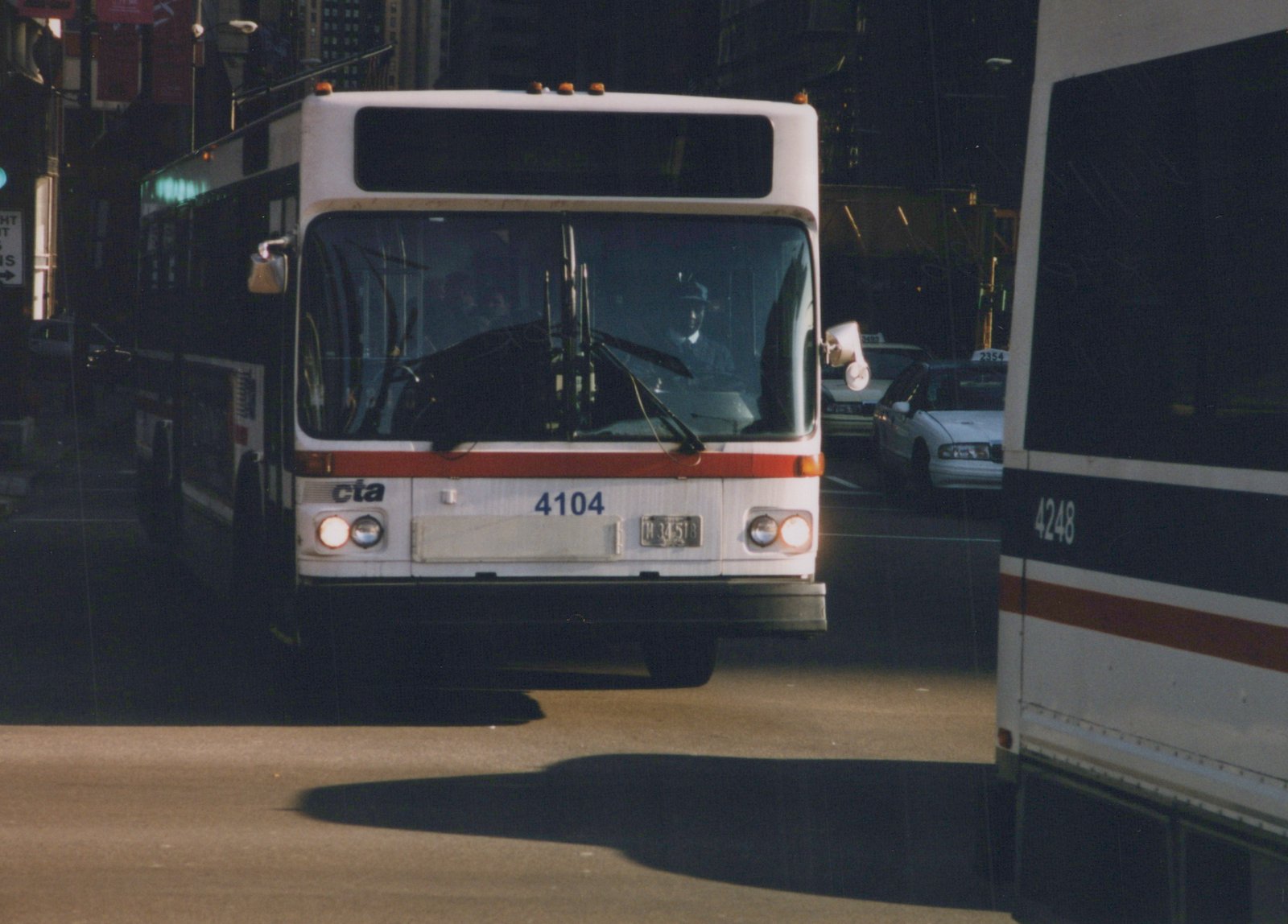 Front view of a white and red Chicago Transit Authority bus, number 4104, with a driver visible through the windshield, parked or moving slowly on a city street next to another CTA bus, number 4248, and some taxis in the background.