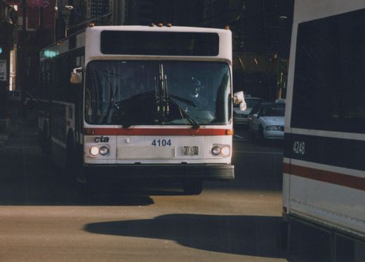 Front view of a white and red Chicago Transit Authority bus, number 4104, with a driver visible through the windshield, parked or moving slowly on a city street next to another CTA bus, number 4248, and some taxis in the background.