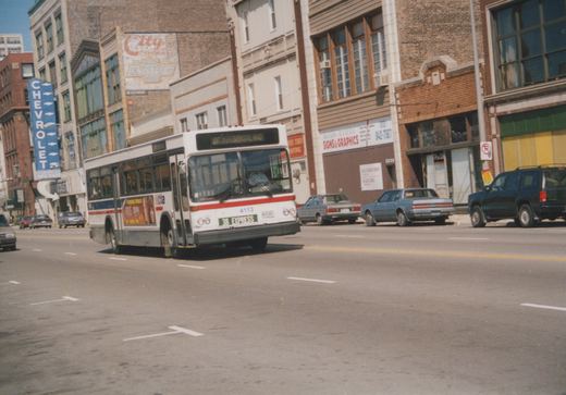A white, red, and blue CTA bus labeled '38 EXPRESS' drives down a city street lined with brick buildings and parked cars on a sunny day.