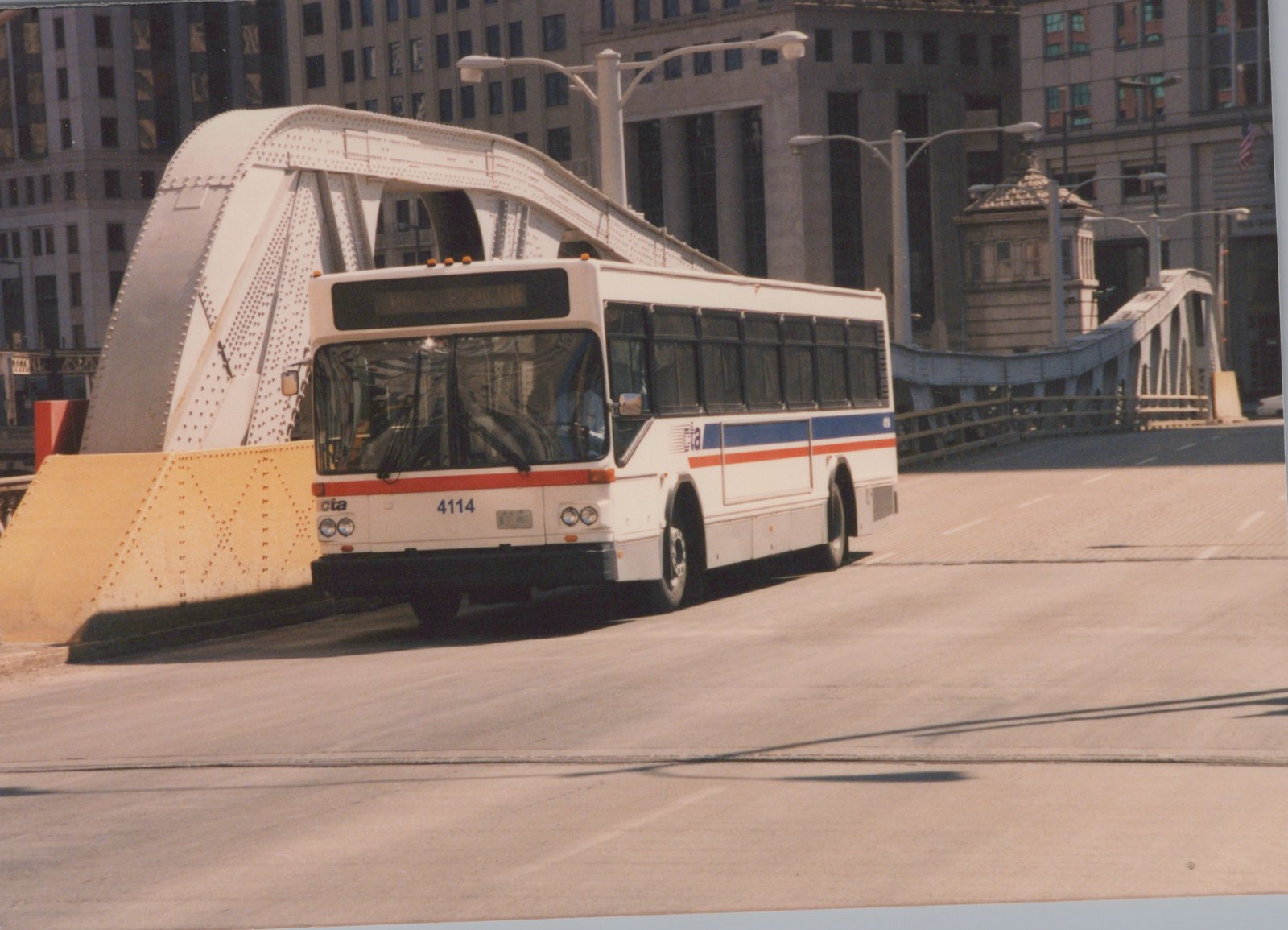 A white CTA bus with blue and red stripes, numbered 4114, is driving on a street across a white steel truss bridge with large buildings in the background.