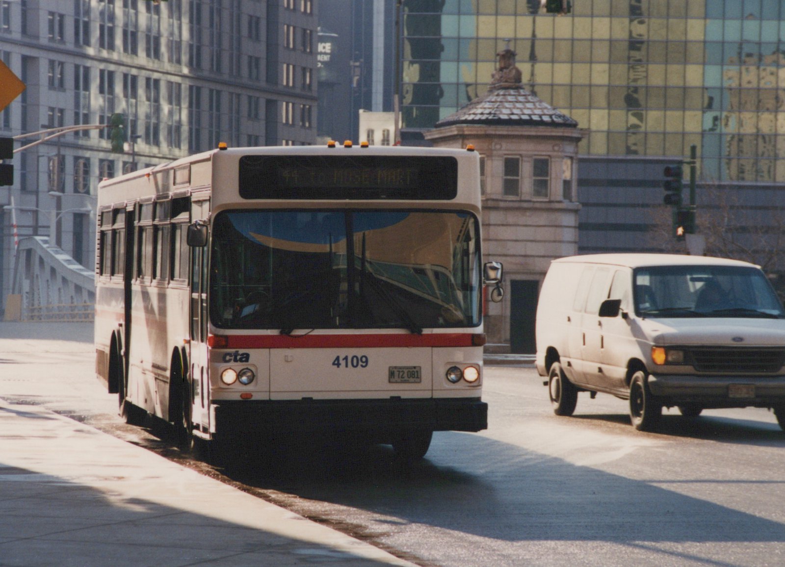 A white and red Chicago Transit Authority bus, number 4109, is driving on a city street next to a white van, with modern buildings and a bridge in the background.