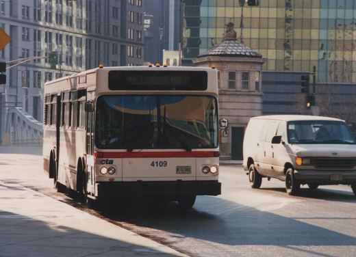 A white and red Chicago Transit Authority bus, number 4109, is driving on a city street next to a white van, with modern buildings and a bridge in the background.