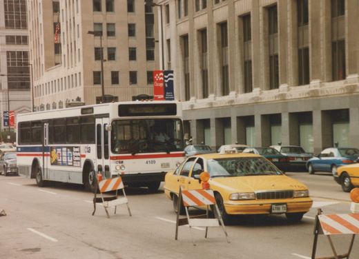 A white CTA bus with the number 4109 on its front, stopped on a city street next to a yellow taxi and orange-and-white barricades, with a large building and 'Chicago '96' banners in the background.