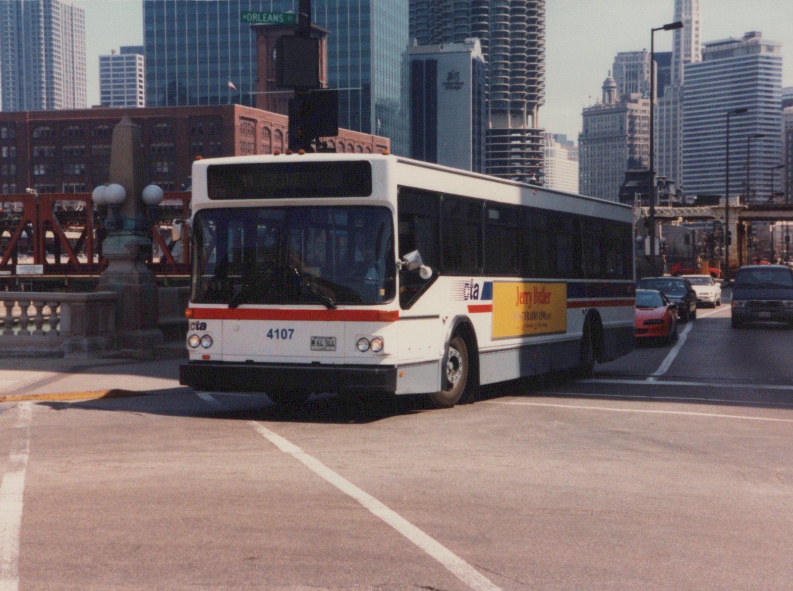 A white CTA bus with a red stripe and the number 4107 is driving on a street in a city with tall buildings behind it.