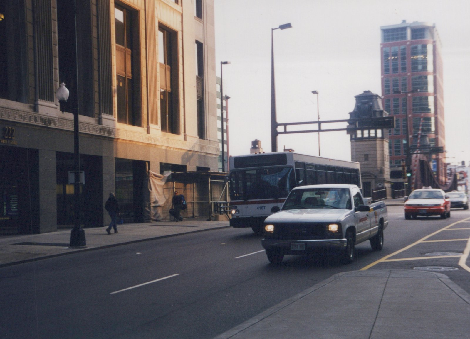 A white and blue Chicago Transit Authority bus drives down a city street, with pedestrians on the sidewalk and a white pickup truck in the foreground.