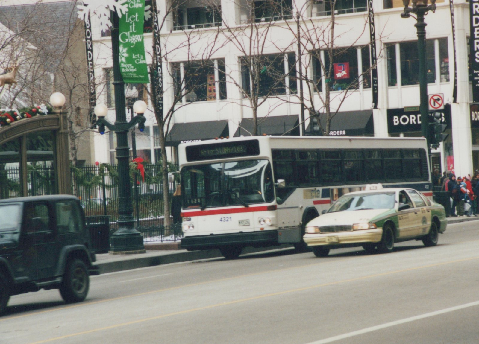 A white and red Chicago Transit Authority bus, numbered 4321, is on a city street during winter, alongside a green and white taxi and a dark SUV, with holiday banners on the lampposts and a 'BORDERS' bookstore in the background.