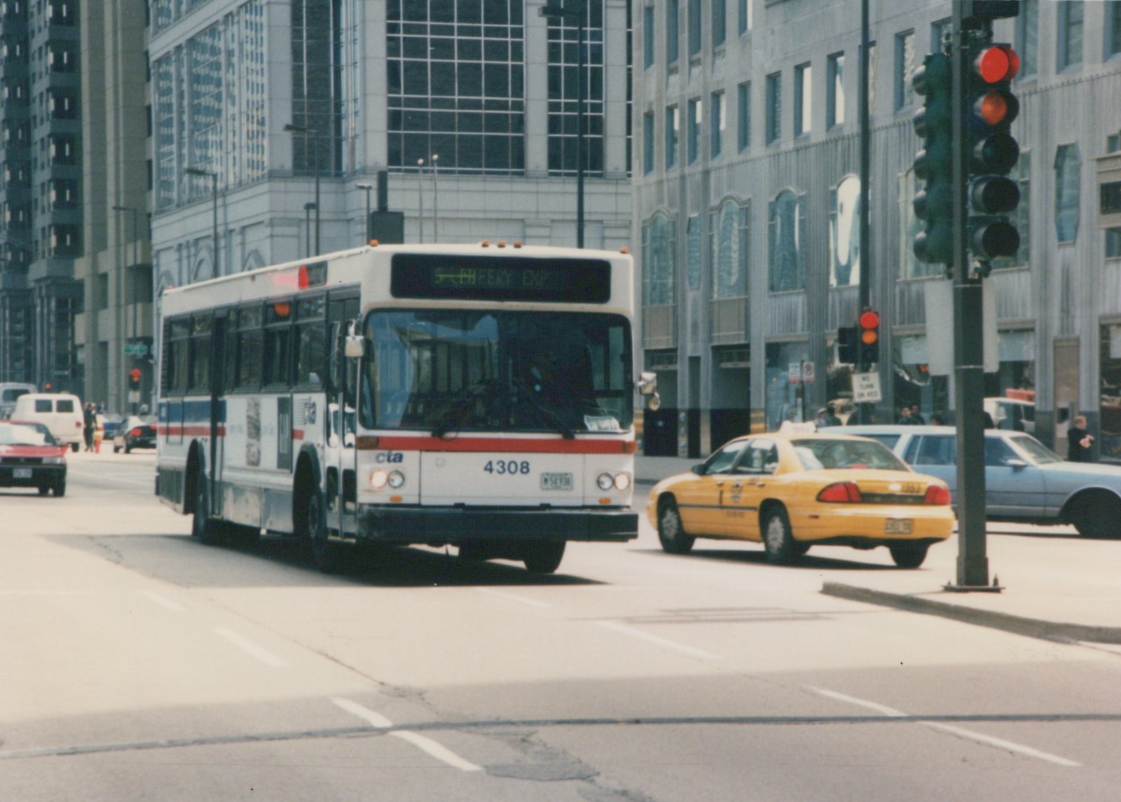 Front three-quarter view of a white CTA bus with fleet number 4308 on a city street, next to a yellow taxi, with tall buildings in the background.