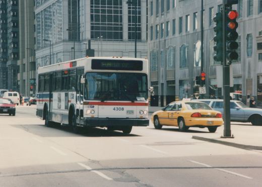 Front three-quarter view of a white CTA bus with fleet number 4308 on a city street, next to a yellow taxi, with tall buildings in the background.