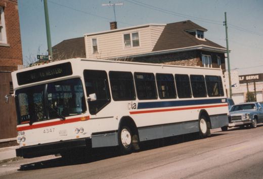 A white, blue, and red Chicago Transit Authority bus with the number 4347 is stopped on a city street in front of a two-story house, with a green street sign for East 87th Street visible.