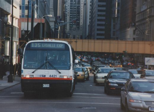 Front view of a white and red Chicago Transit Authority bus with the number 4421, showing route 135 LaSalle Express on its sign, driving on a busy city street with an elevated train track overhead.