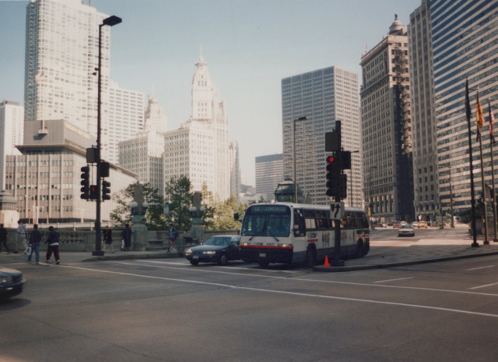 A white and blue CTA bus, number 4421, is stopped at a city intersection with tall buildings in the background and pedestrians on the sidewalk.