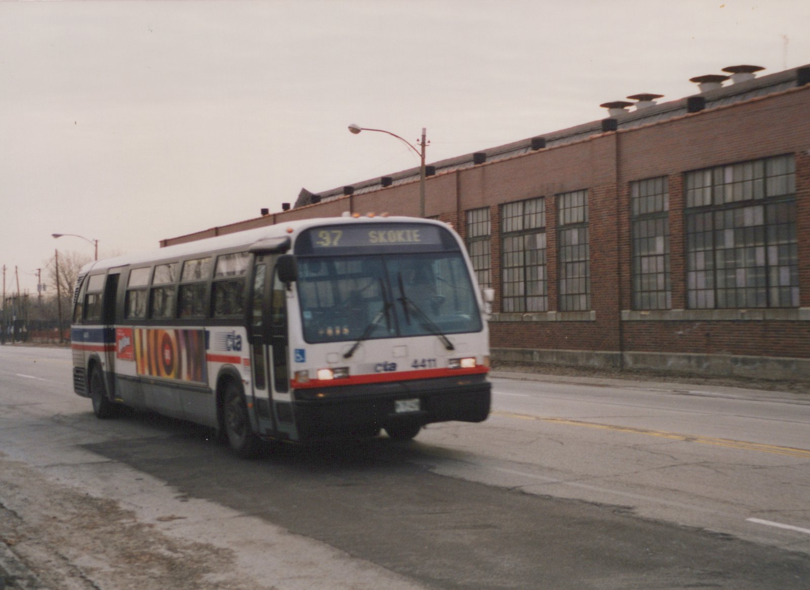A white Chicago Transit Authority motor bus numbered 4411 is driving down a street in front of a large brick building on an overcast winter day.