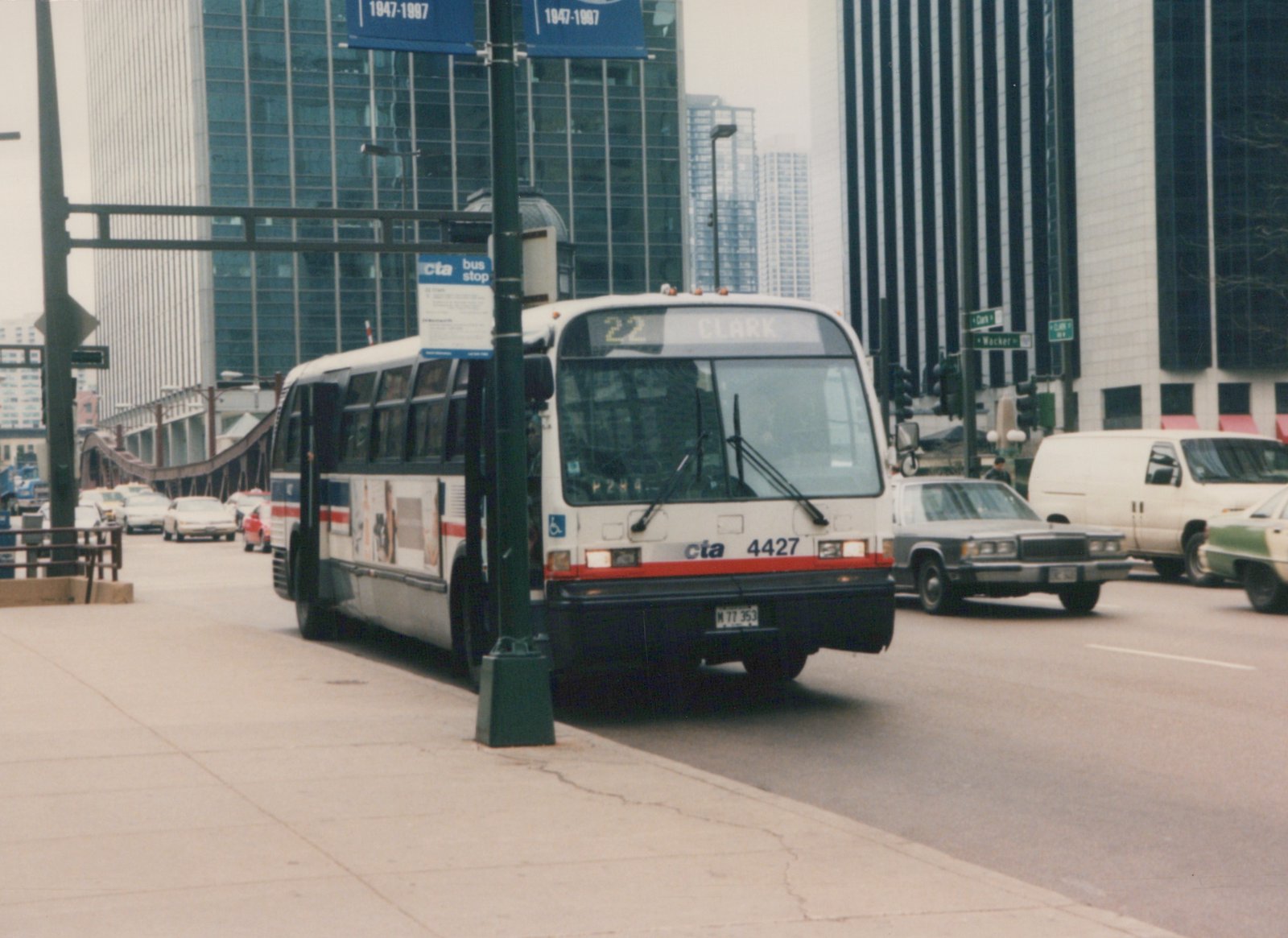 A white Chicago Transit Authority bus with the number 4427 is stopped on a city street at an intersection with street signs for Clark Street and Wacker Drive, surrounded by tall buildings and traffic.