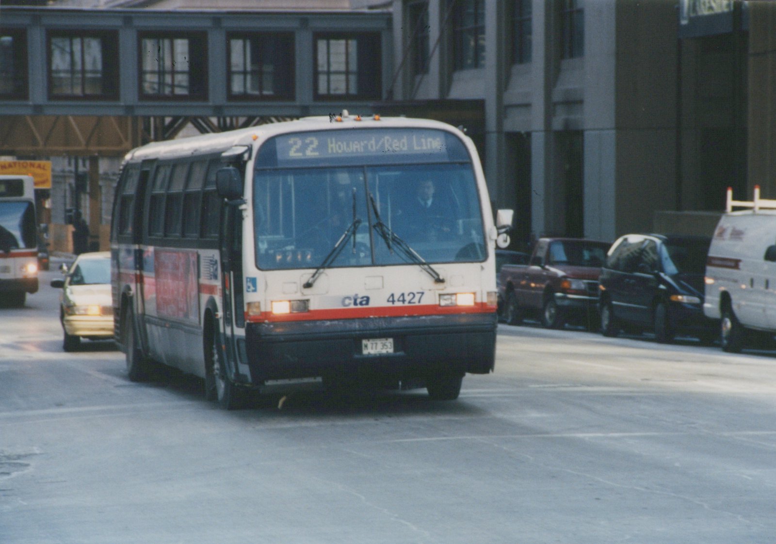 Front three-quarter view of a white Chicago Transit Authority bus number 4427 with red and blue stripes, displaying route 22 to Howard/Red Line, on a city street with an elevated structure overhead and other vehicles nearby.