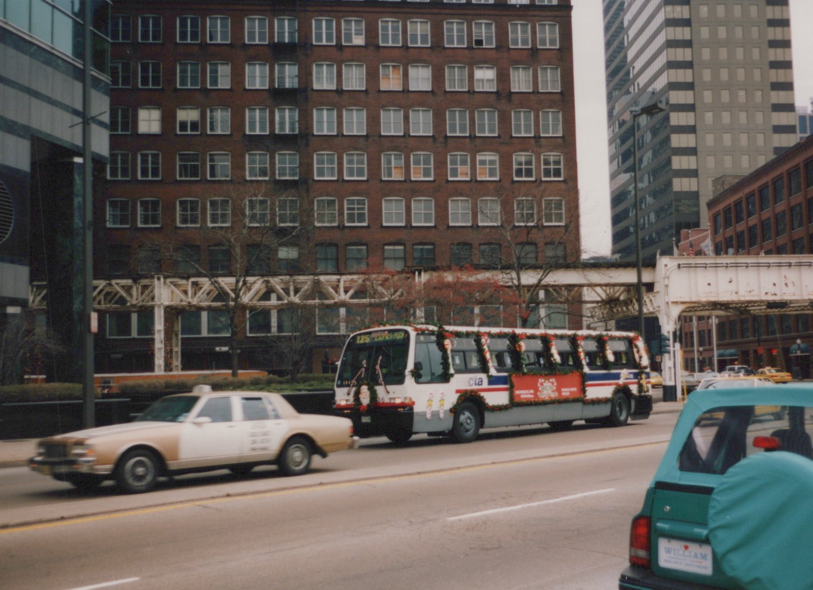 A white Chicago Transit Authority bus with holiday decorations is driving on a city street, with a beige taxi and a teal car nearby. Tall buildings and a steel bridge are in the background.