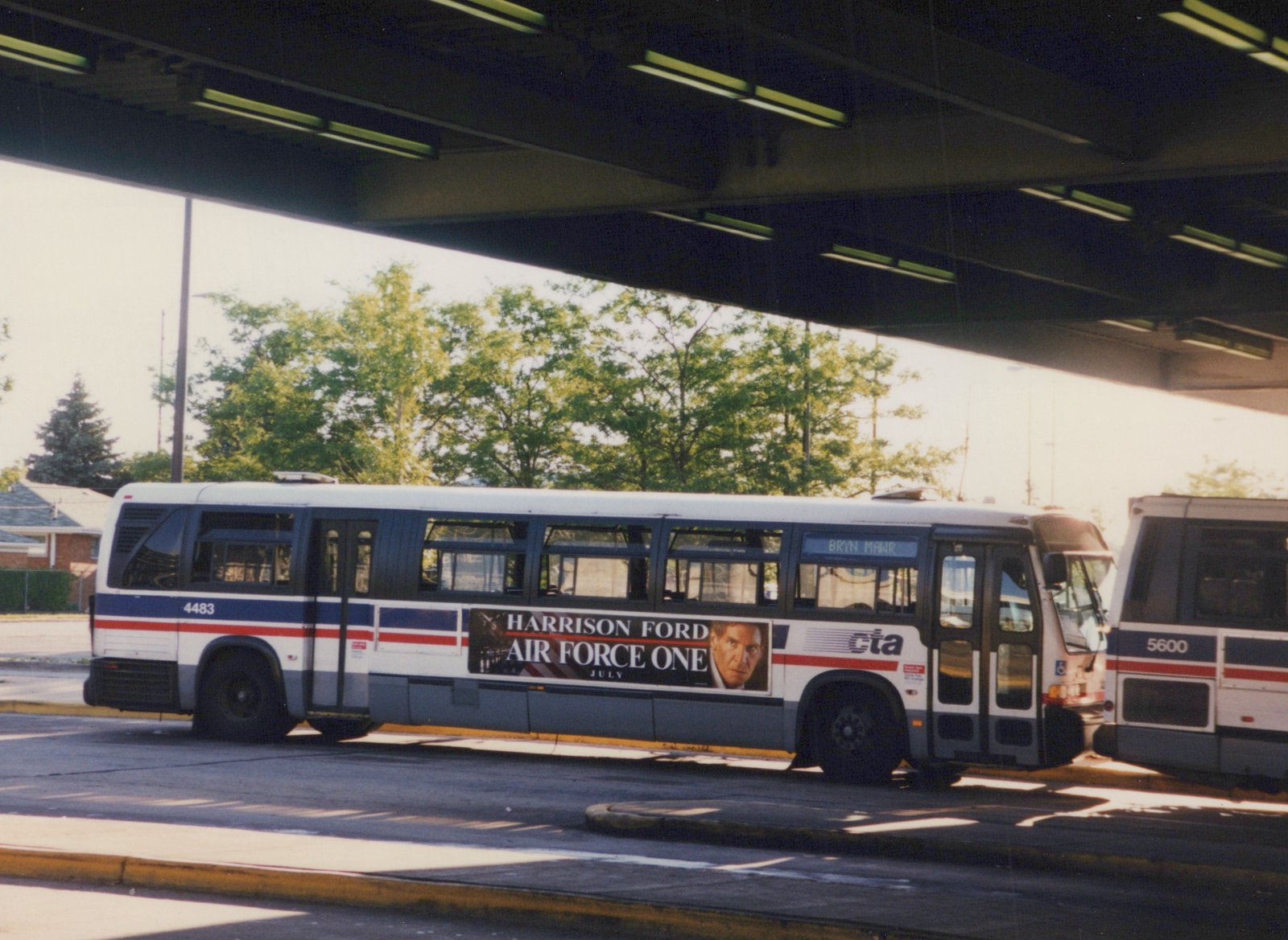 A white and blue CTA bus displaying a Harrison Ford 'Air Force One' movie ad, parked under a highway overpass with green trees in the background.