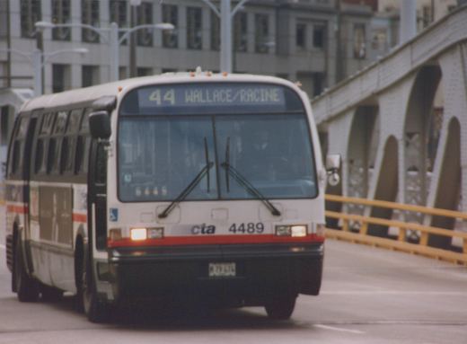 A white, red, and gray CTA bus, number 4489, on route 44 Wallace/Racine, drives across a bridge in a city setting.