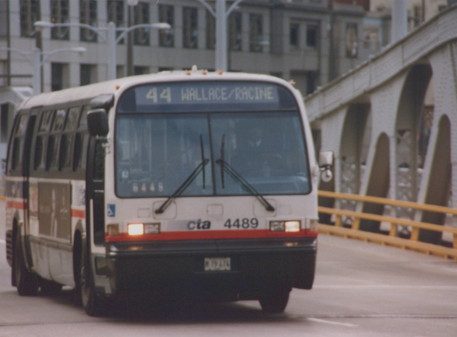 A white, red, and gray CTA bus, number 4489, on route 44 Wallace/Racine, drives across a bridge in a city setting.