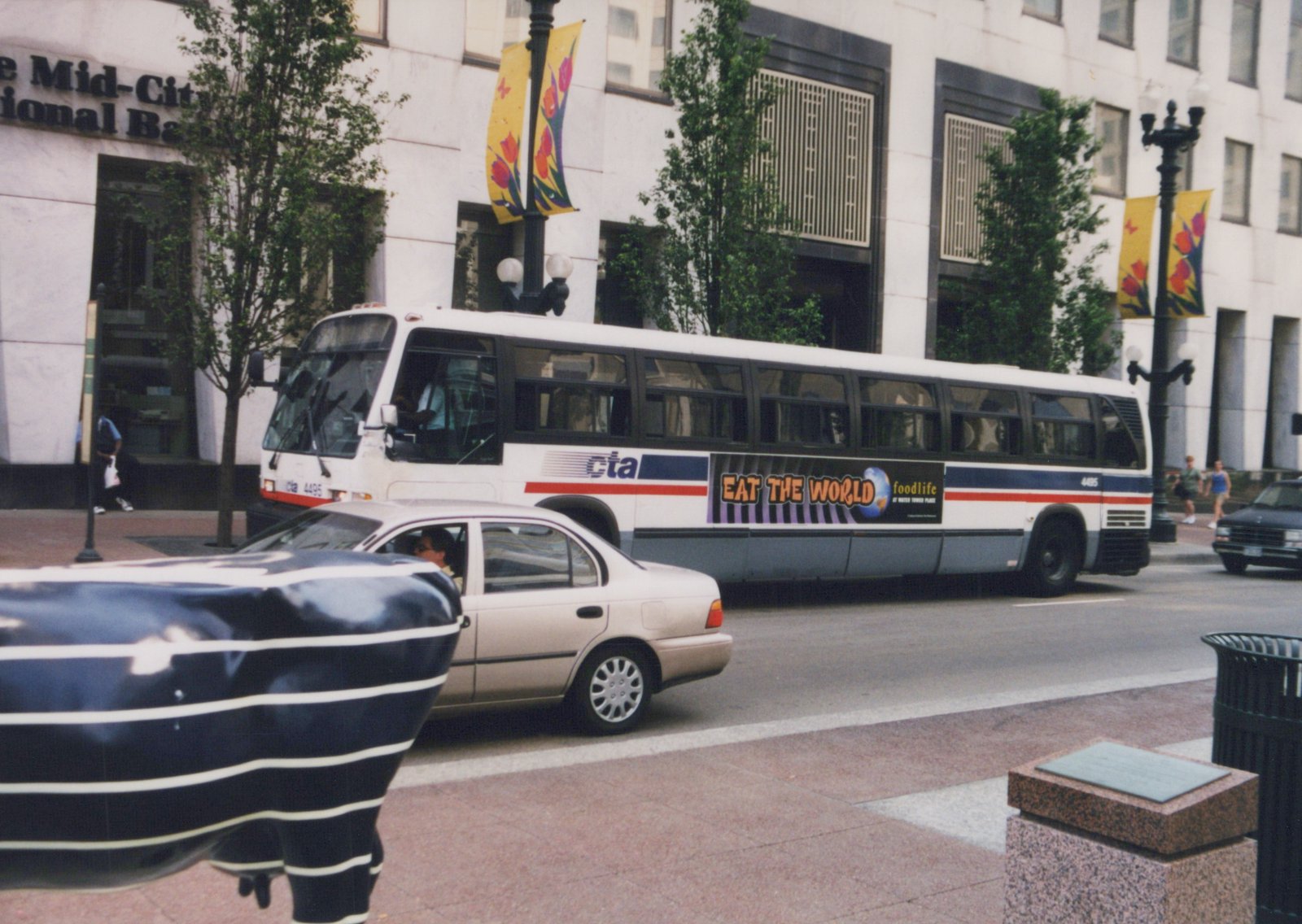 A white CTA bus numbered 4495 with a blue and red stripe runs down a city street lined with trees and a large building, with a light-colored car in the foreground and pedestrians on the sidewalk.