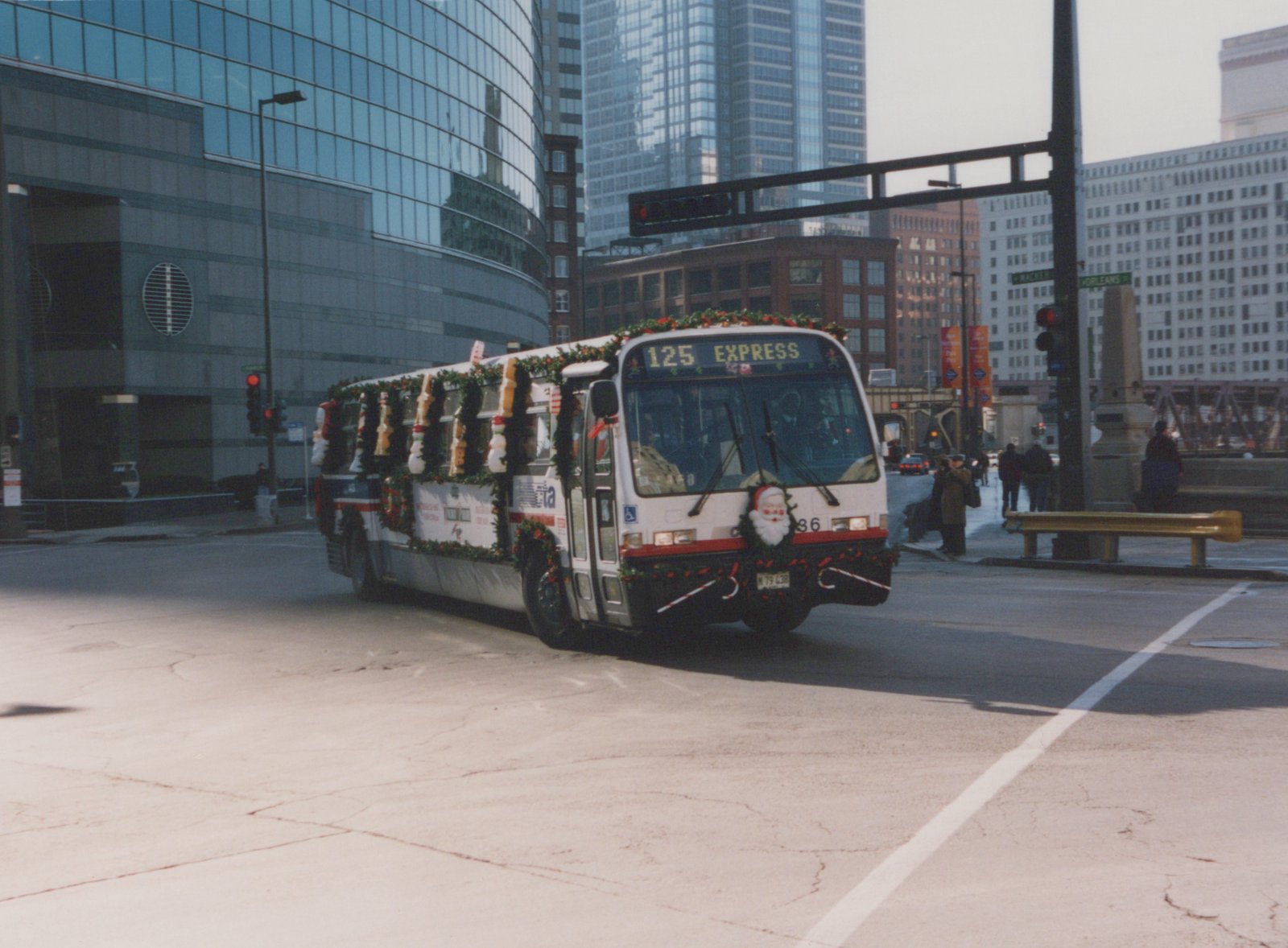 A white Chicago Transit Authority bus decorated with Christmas garlands and a Santa figure on the front, driving on a city street with modern buildings in the background.